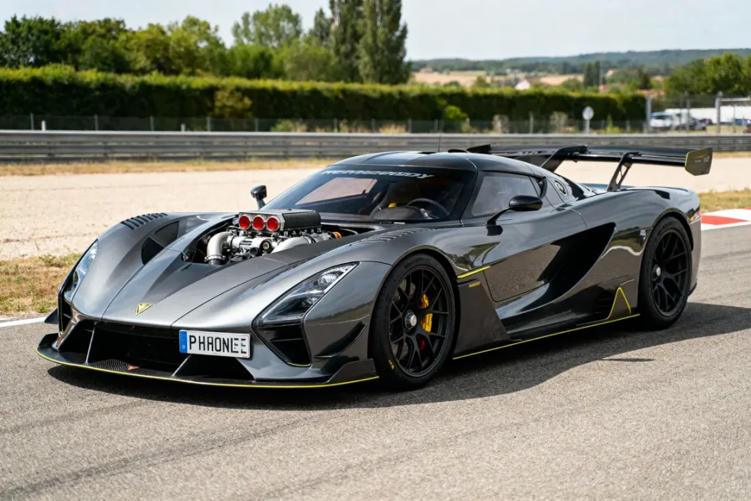 Voiture de sport grise sur circuit, moteur visible, aileron arrière, arrière-plan verdoyant.