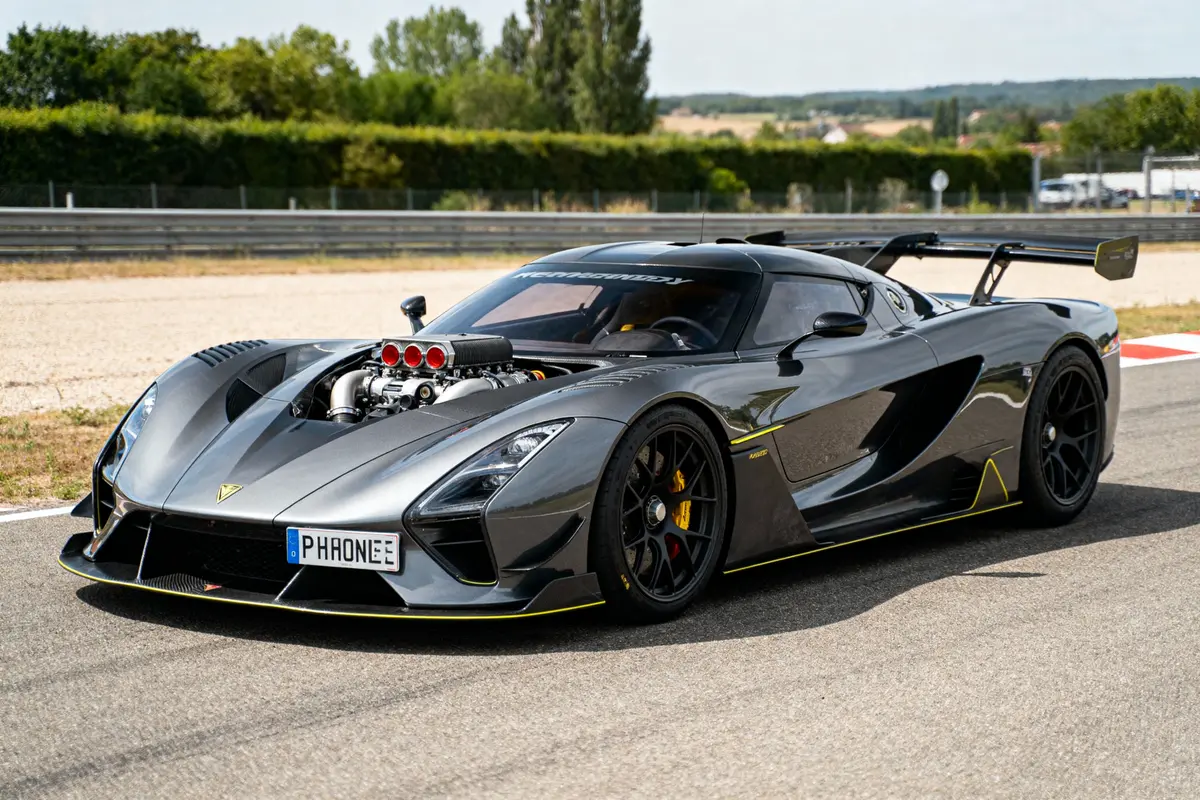Voiture de sport grise sur circuit, moteur visible, aileron arrière, arrière-plan verdoyant.