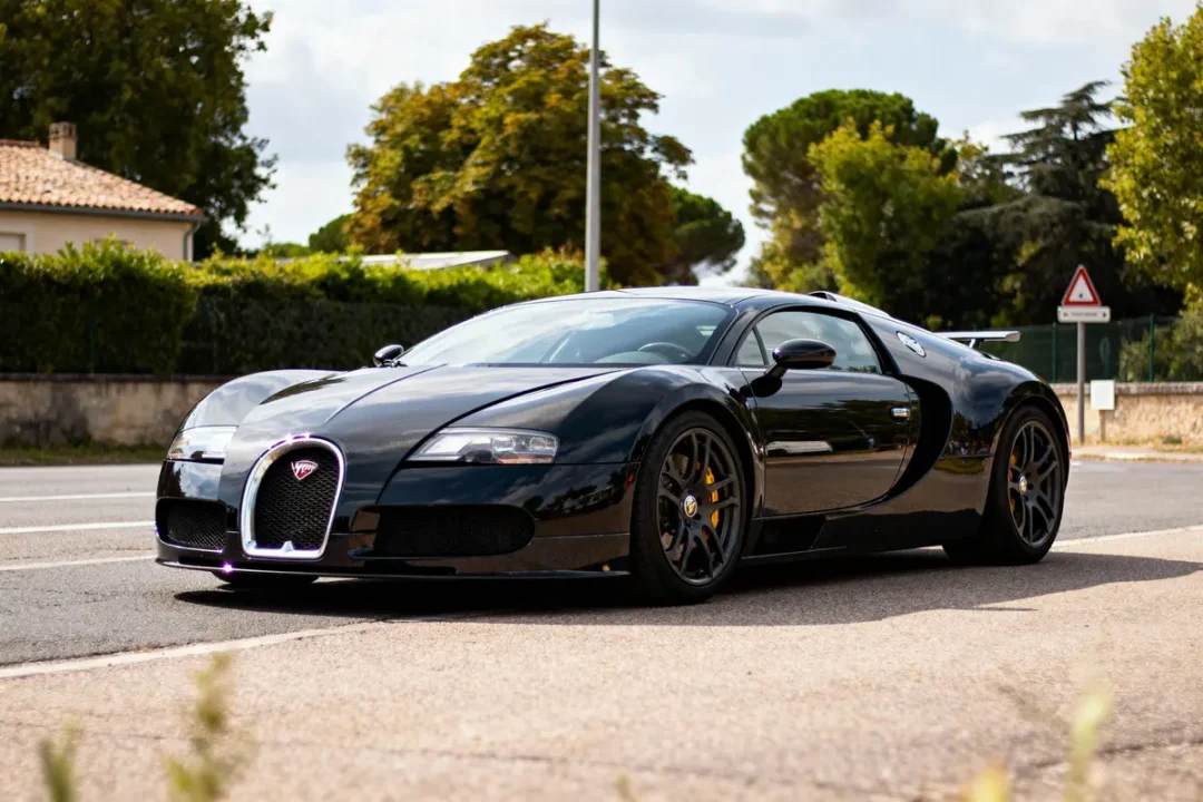 Voiture de sport noire Bugatti sur route, entourée d'arbres et de maisons. Temps ensoleillé.