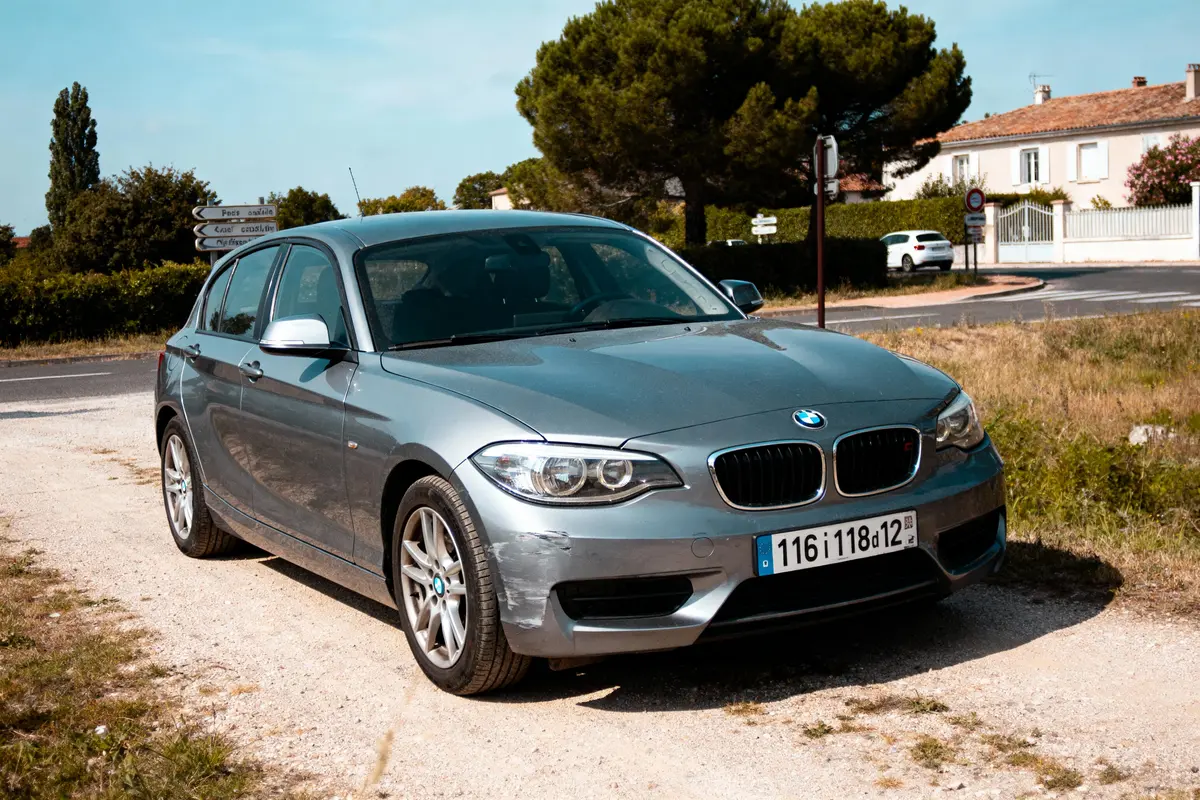 Voiture BMW grise garée sur un chemin de terre, avec végétation et maisons en arrière-plan.