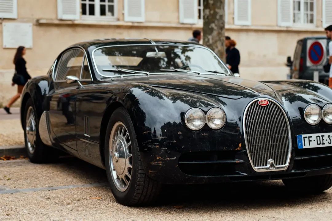 Voiture noire classique garée, avec phares ronds et calandre vintage, devant bâtiment avec fenêtres.
