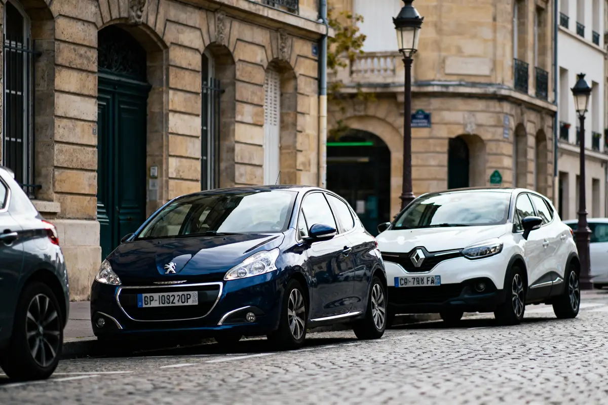 Deux voitures, Peugeot bleue et Renault blanche, stationnées dans une rue pavée parisienne, bâtiments historiques en arrière-plan.