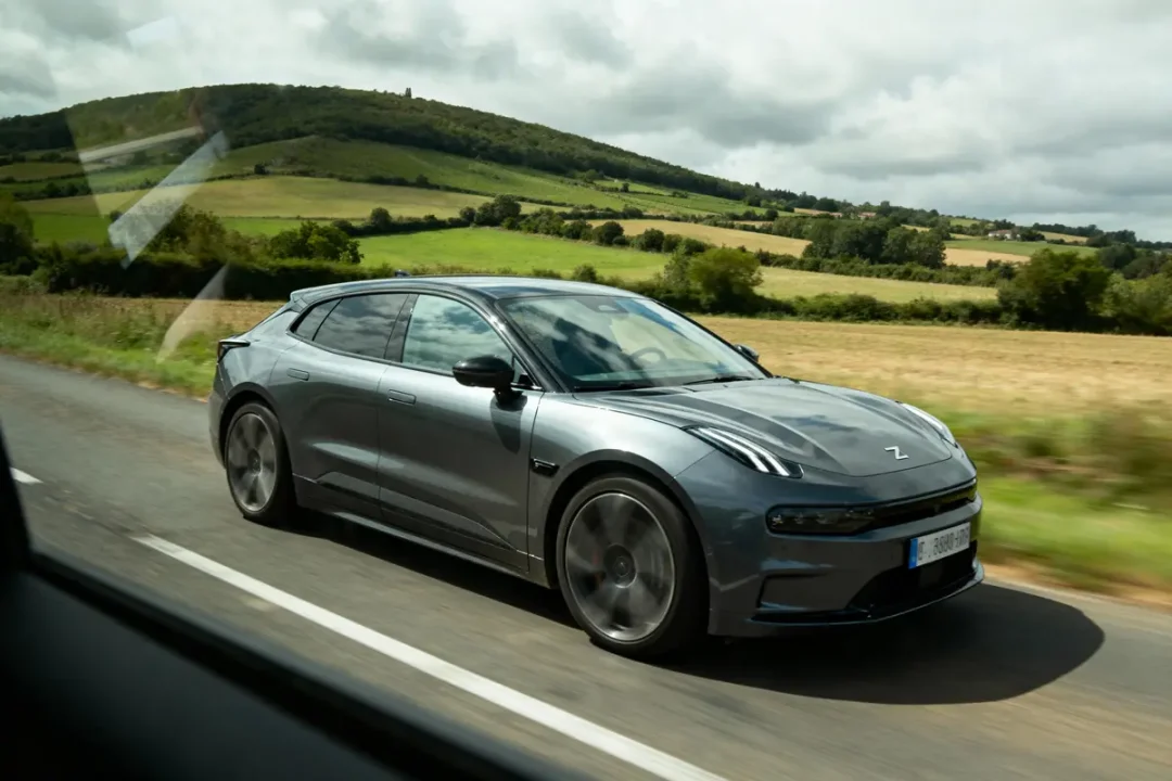 Voiture électrique grise sur route de campagne, collines et champs en arrière-plan, ciel nuageux.