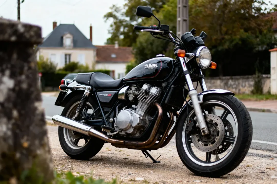 Moto Yamaha noire, garée en bord de route, maison floue en arrière-plan, arbres et ciel nuageux.