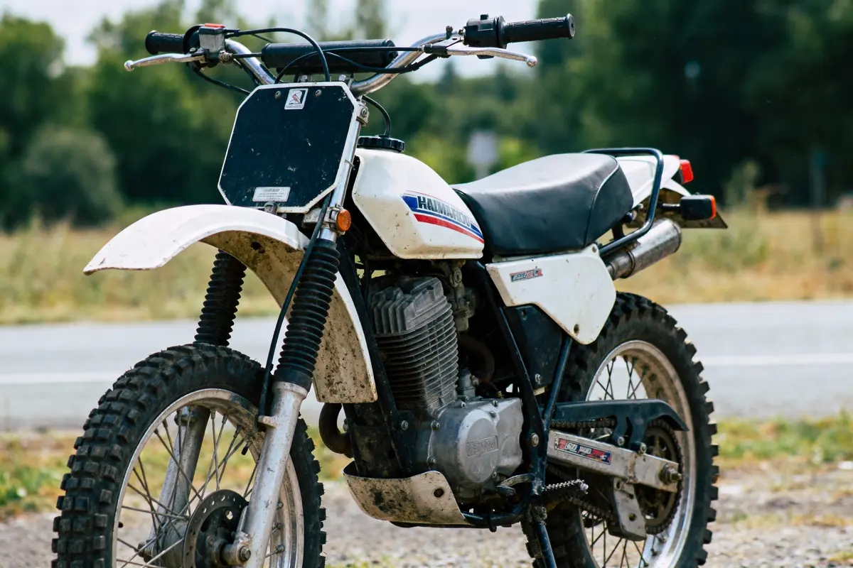 Moto tout-terrain vintage blanche, pose en extérieur, pneus avec crampons, fond de nature floue.
