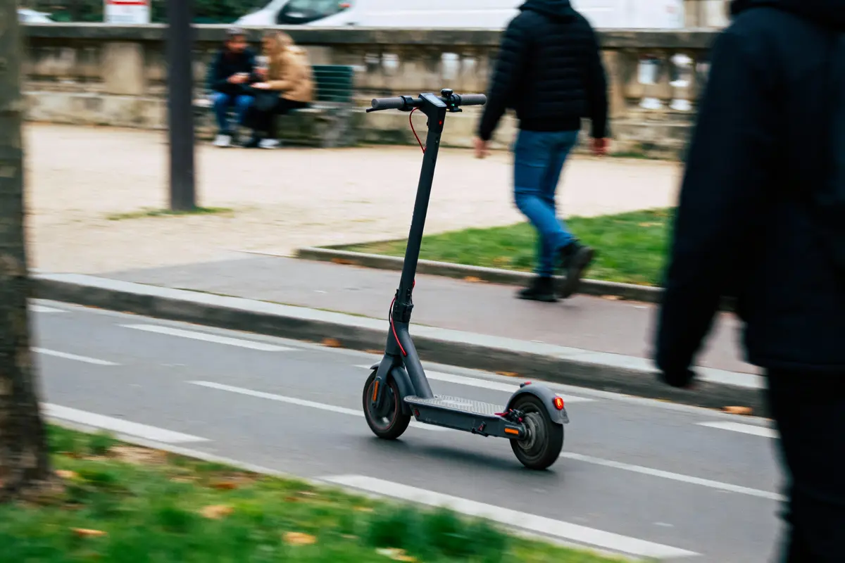 Trottinette électrique noire sur une piste cyclable, personnes floues et bancs en arrière-plan.