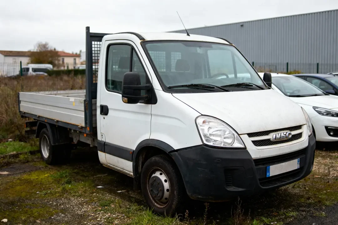 Camion blanc utilitaire garé sur terrain herbeux, temps nuageux, hangar en arrière-plan.