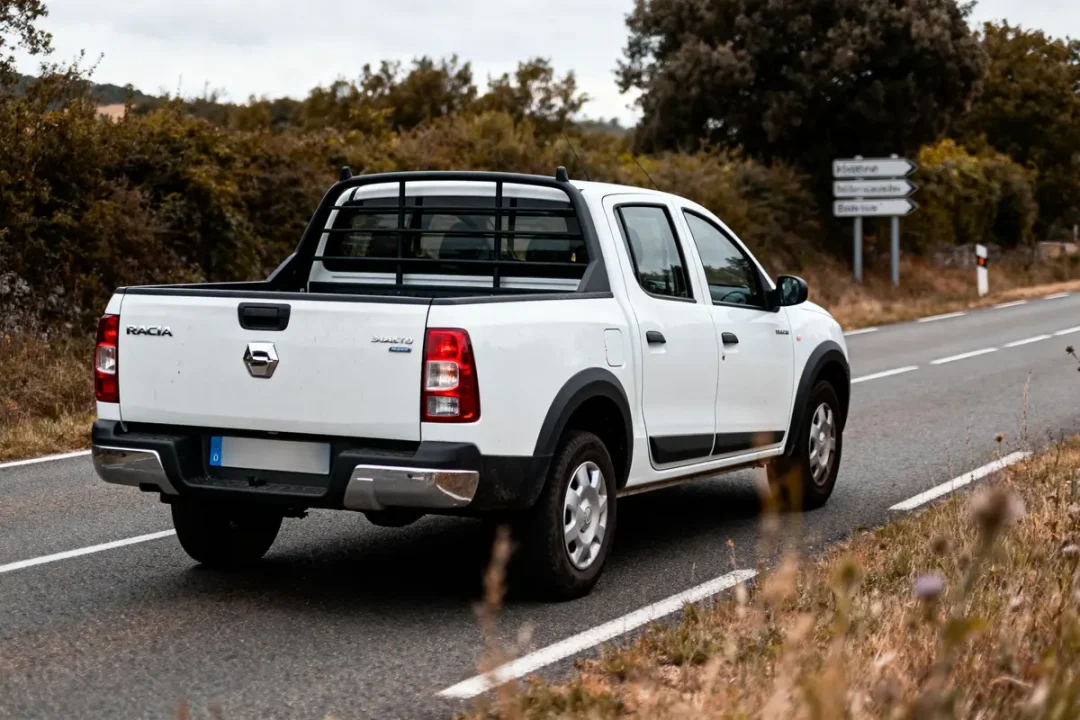 Pick-up blanc circulant sur une route de campagne, entouré de végétation et de panneaux de signalisation en arrière-plan.