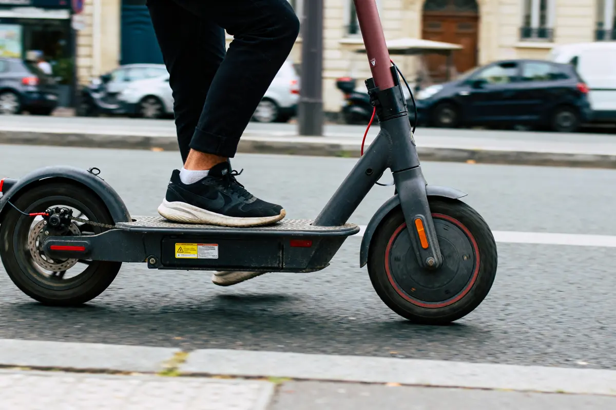 Personne conduisant une trottinette électrique noire sur route en ville, portant des chaussures noires et blanc.