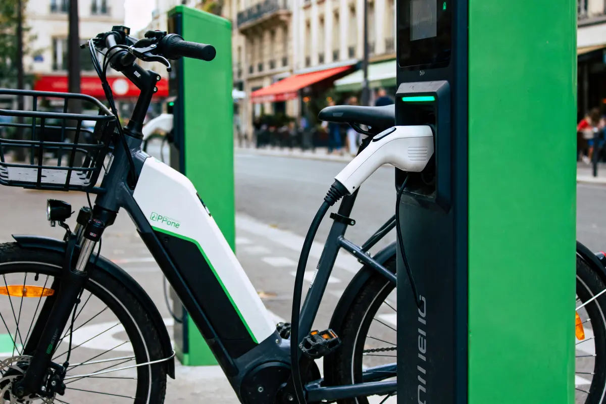 Vélo électrique branché à une borne de recharge sur un trottoir urbain, avec bâtiments en arrière-plan.