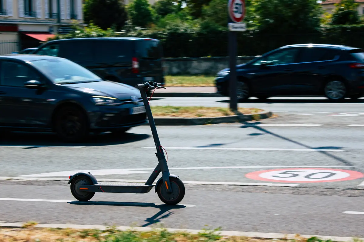 Trottinette électrique noire sur piste cyclable, voitures en arrière-plan, panneau de vitesse 50 km/h visible.