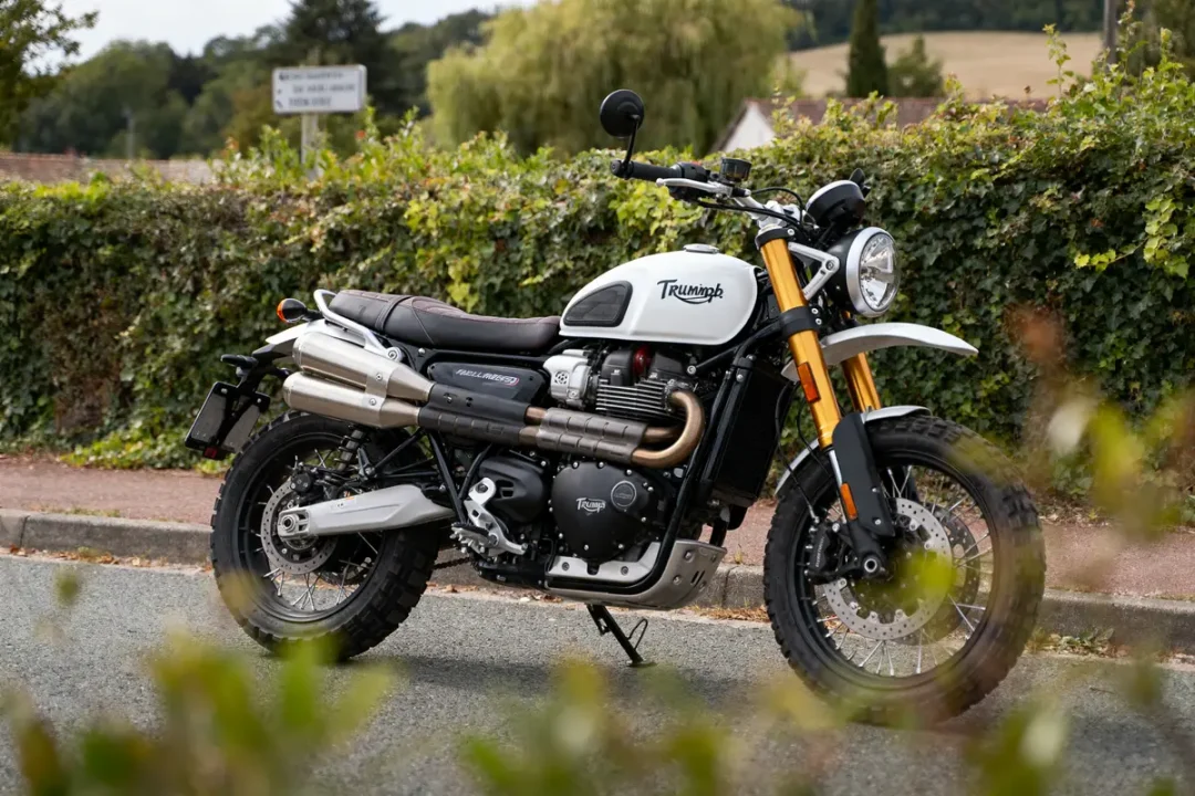 Moto Triumph blanc et noir garée sur une route, végétation en arrière-plan.