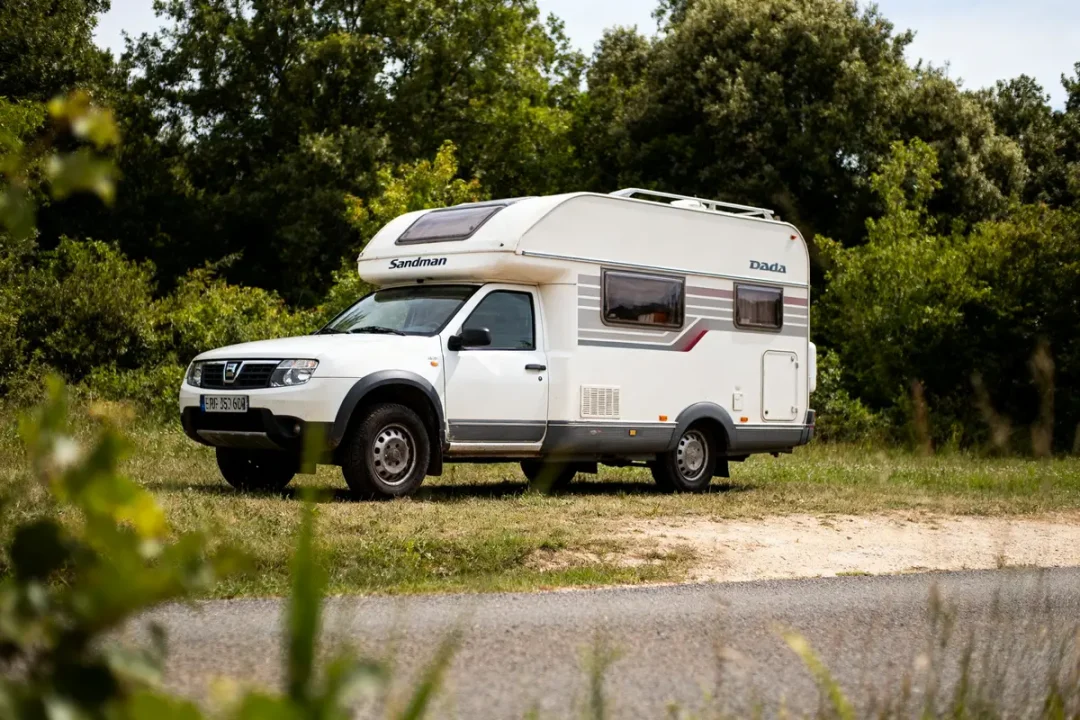 Camping-car blanc stationné dans la nature, entouré d'arbres et de verdure en plein air, sur chemin.