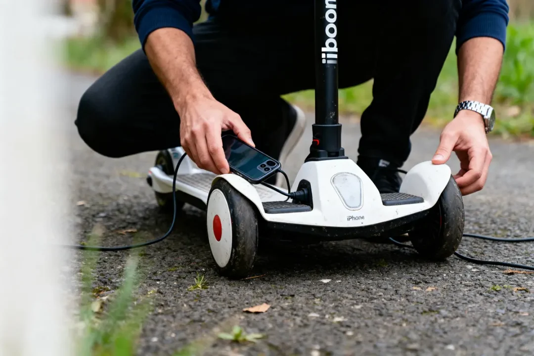 Personne accroupie charge une trottinette électrique blanche, tenant un smartphone, sur chemin asphalté extérieur.