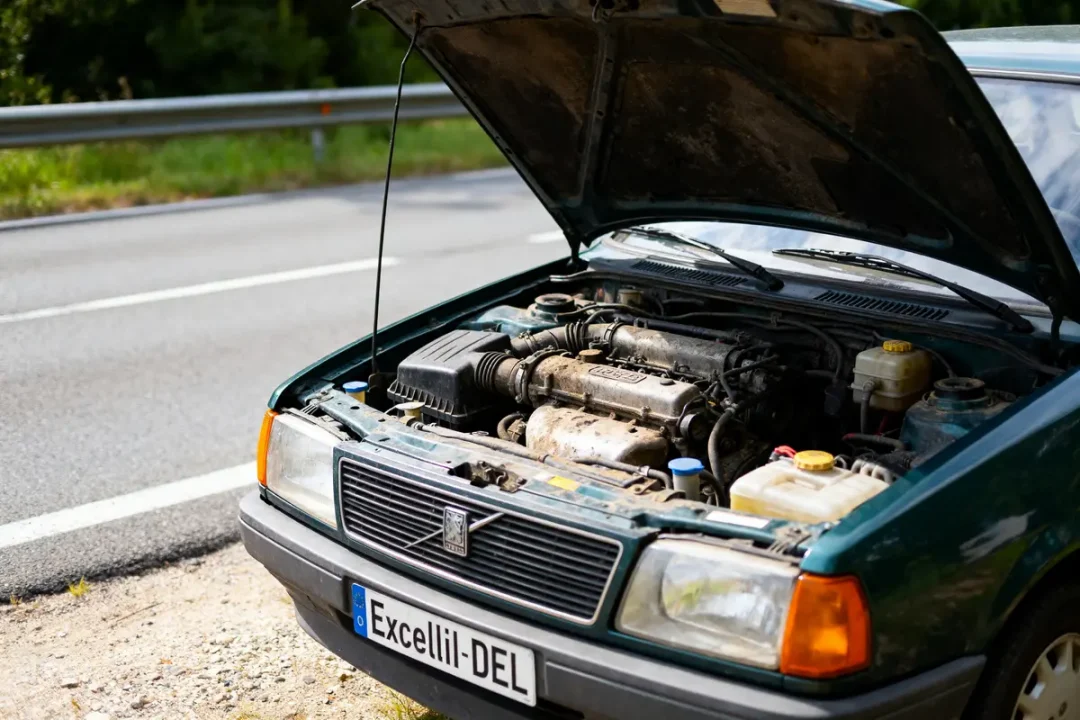 Voiture verte avec capot ouvert sur le bord de la route, moteur visible. Plaque d'immatriculation: "Excellil-DEL".