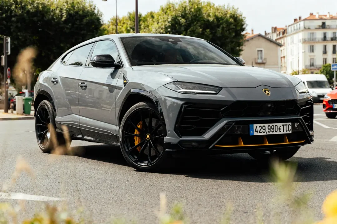 SUV gris Lamborghini sur route urbaine, jantes noires, accents jaunes, bâtiments et arbres en arrière-plan.