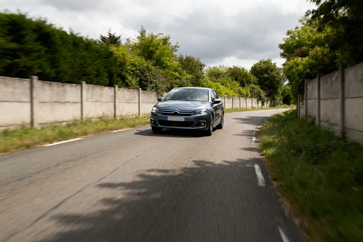 Voiture grise circulant sur une route de campagne bordée d'arbres et de murs en béton.