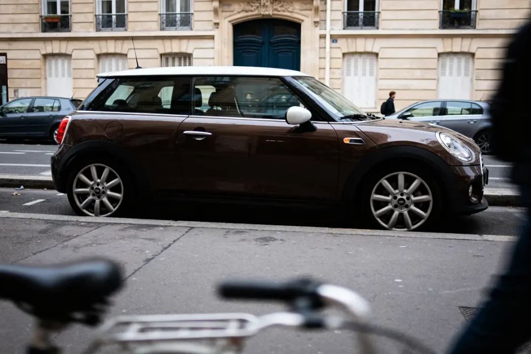 Voiture Mini marron garée, bâtiment parisien en arrière-plan, vélo flou au premier plan.