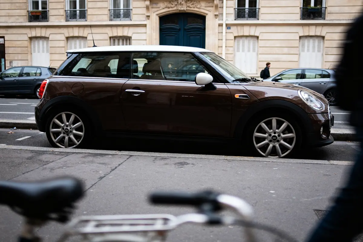 Voiture Mini marron garée, bâtiment parisien en arrière-plan, vélo flou au premier plan.