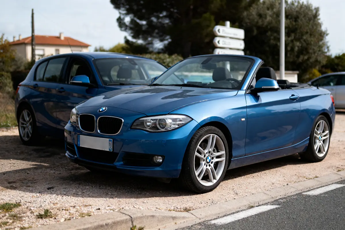 Deux voitures BMW bleues stationnées, l'une coupé cabriolet. Panneaux de signalisation en arrière-plan.