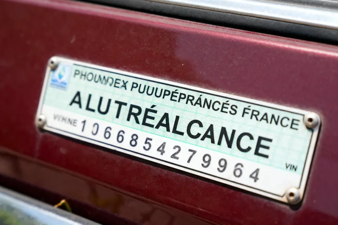 Plaque d’immatriculation humoristique avec texte déformé sur fond de voiture bordeaux. Numéro série en bas.