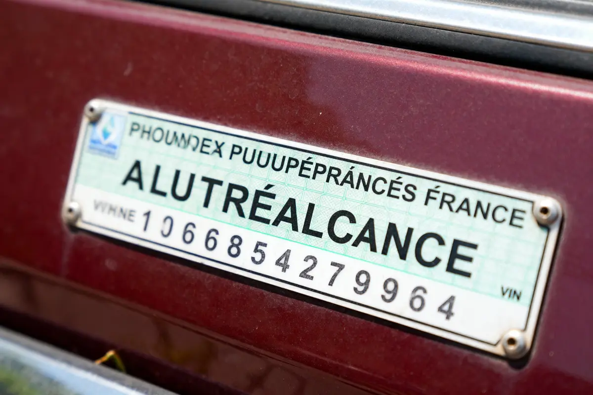 Plaque d’immatriculation humoristique avec texte déformé sur fond de voiture bordeaux. Numéro série en bas.