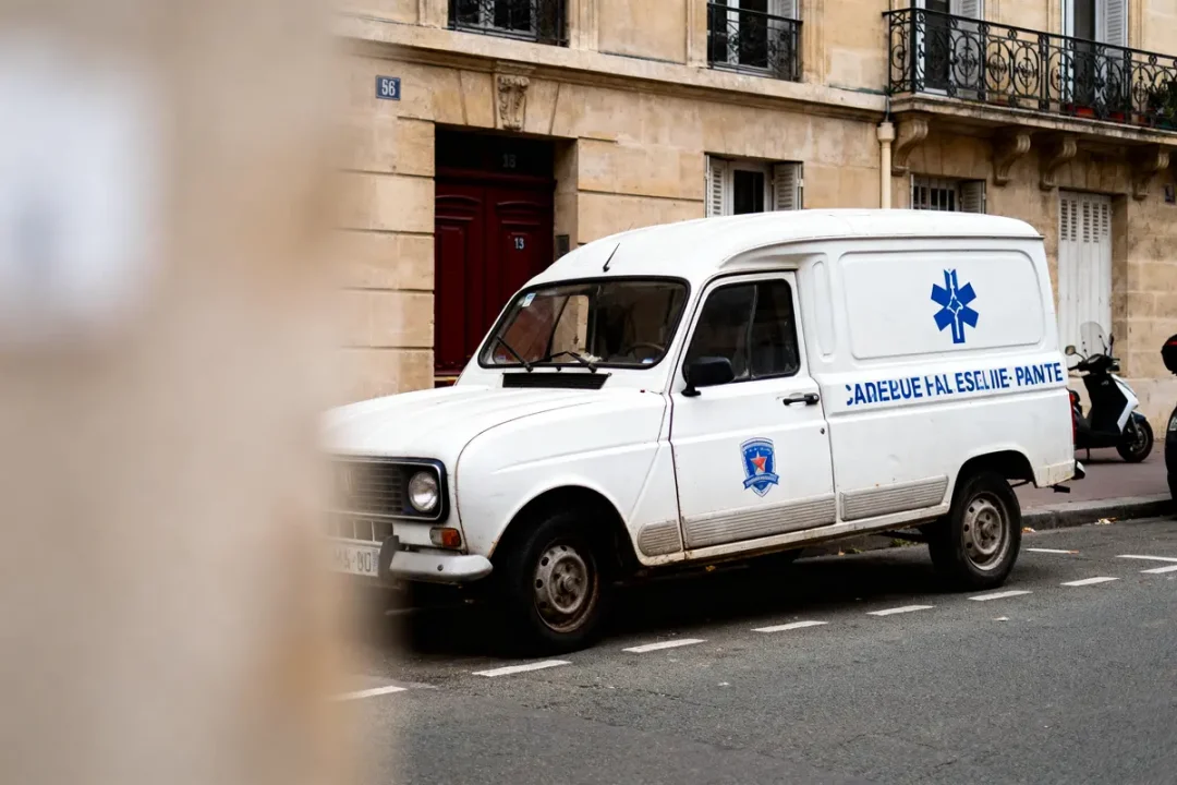 Fourgonnette blanche garée dans la rue, bâtiment en pierre en arrière-plan, texte partiellement lisible sur le véhicule.