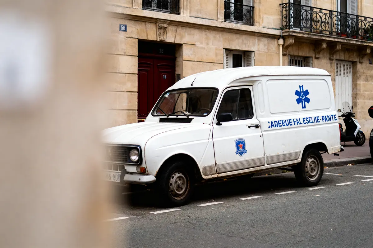 Fourgonnette blanche garée dans la rue, bâtiment en pierre en arrière-plan, texte partiellement lisible sur le véhicule.