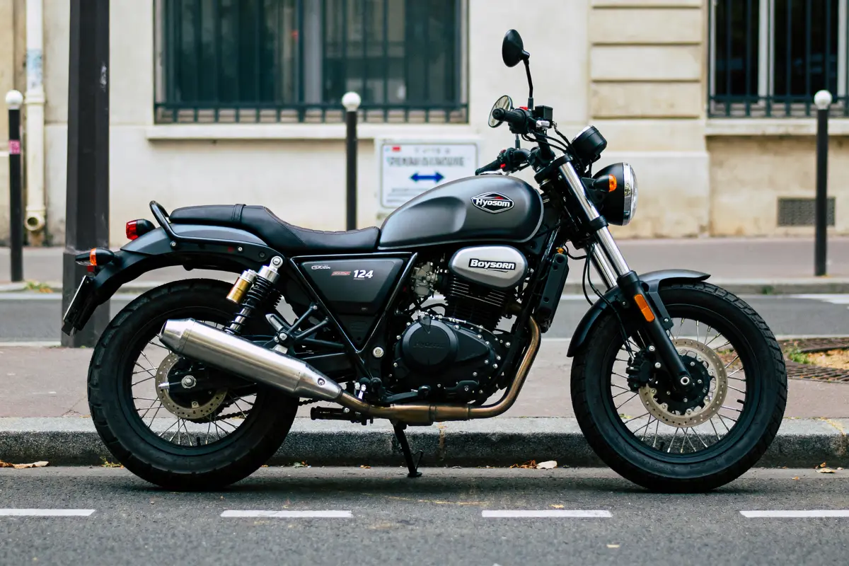Moto noire Hysosn stationnée dans la rue, devant un bâtiment beige. Vue latérale, échappement visible.