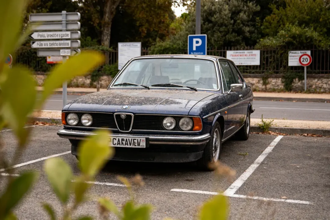 Voiture ancienne BMW stationnée dans un parking, feuillage flou au premier plan, panneaux en arrière-plan.