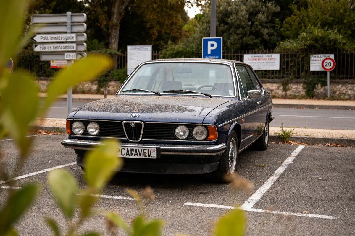 Voiture ancienne BMW stationnée dans un parking, feuillage flou au premier plan, panneaux en arrière-plan.