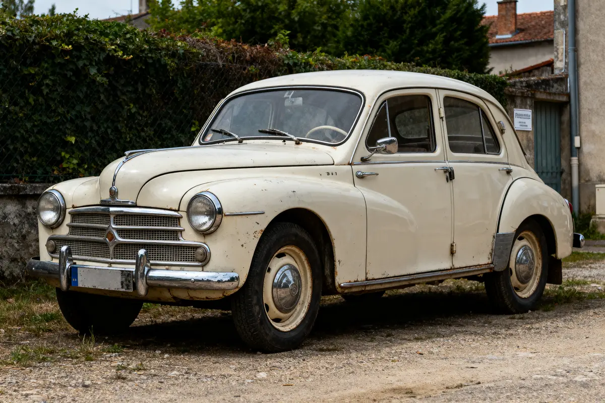 Voiture ancienne blanche garée sur un chemin, entourée de végétation et de bâtiments en arrière-plan.