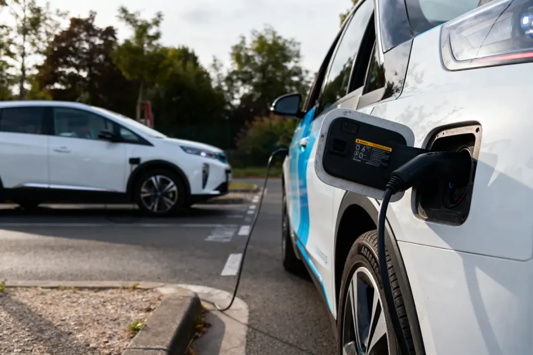 Voiture électrique en charge sur borne, autre véhicule en arrière-plan, stationnement extérieur, arbres environnants.