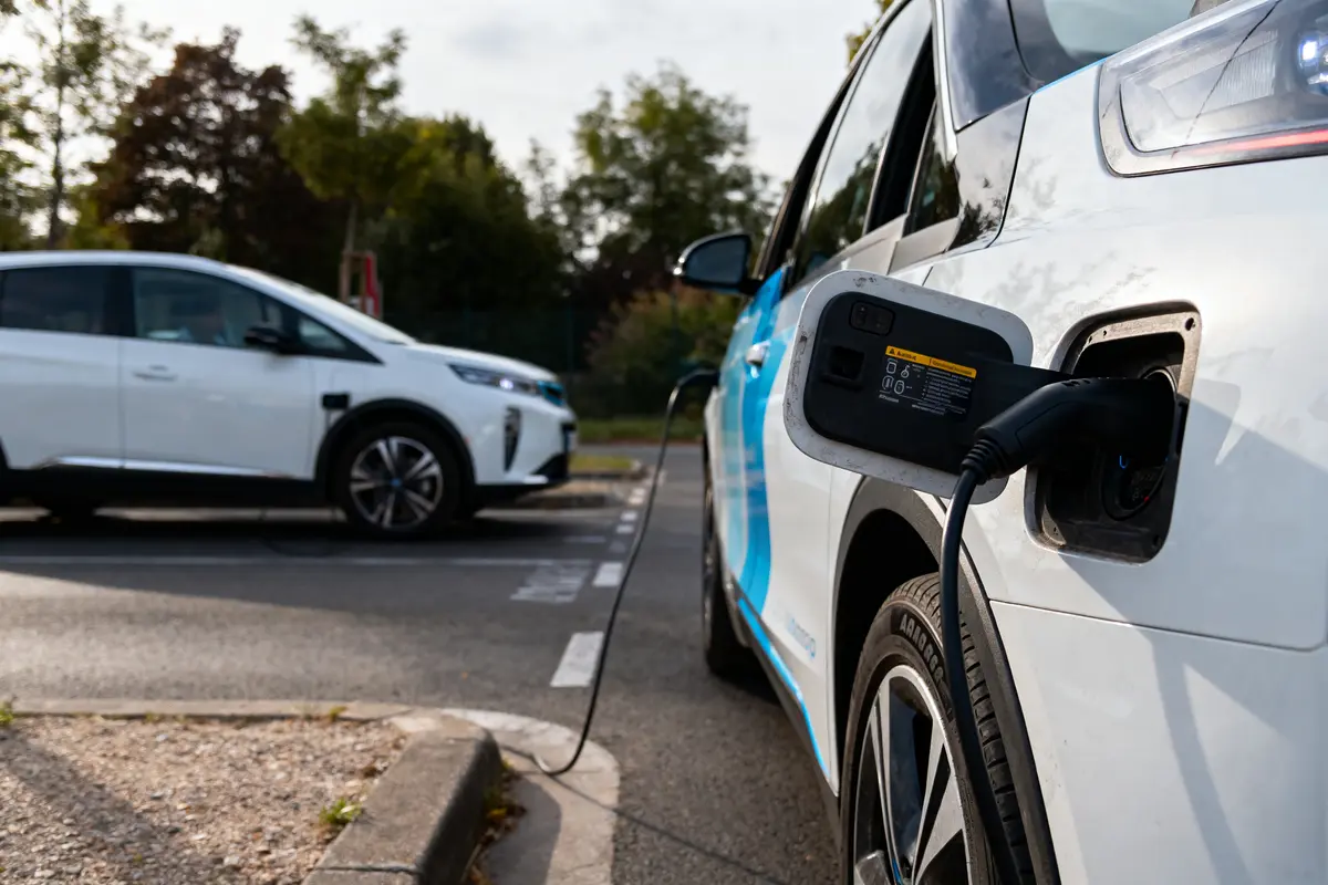 Voiture électrique en charge sur borne, autre véhicule en arrière-plan, stationnement extérieur, arbres environnants.