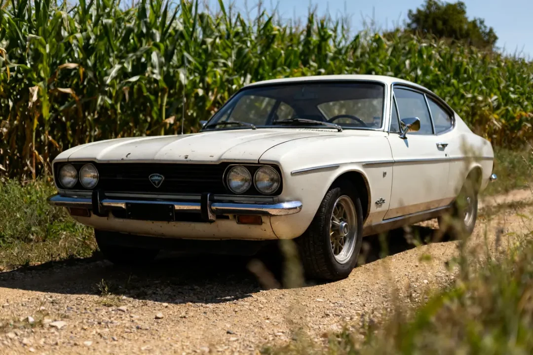 Ford Capri 1970 blanche garée sur chemin de terre près d’un champ de maïs