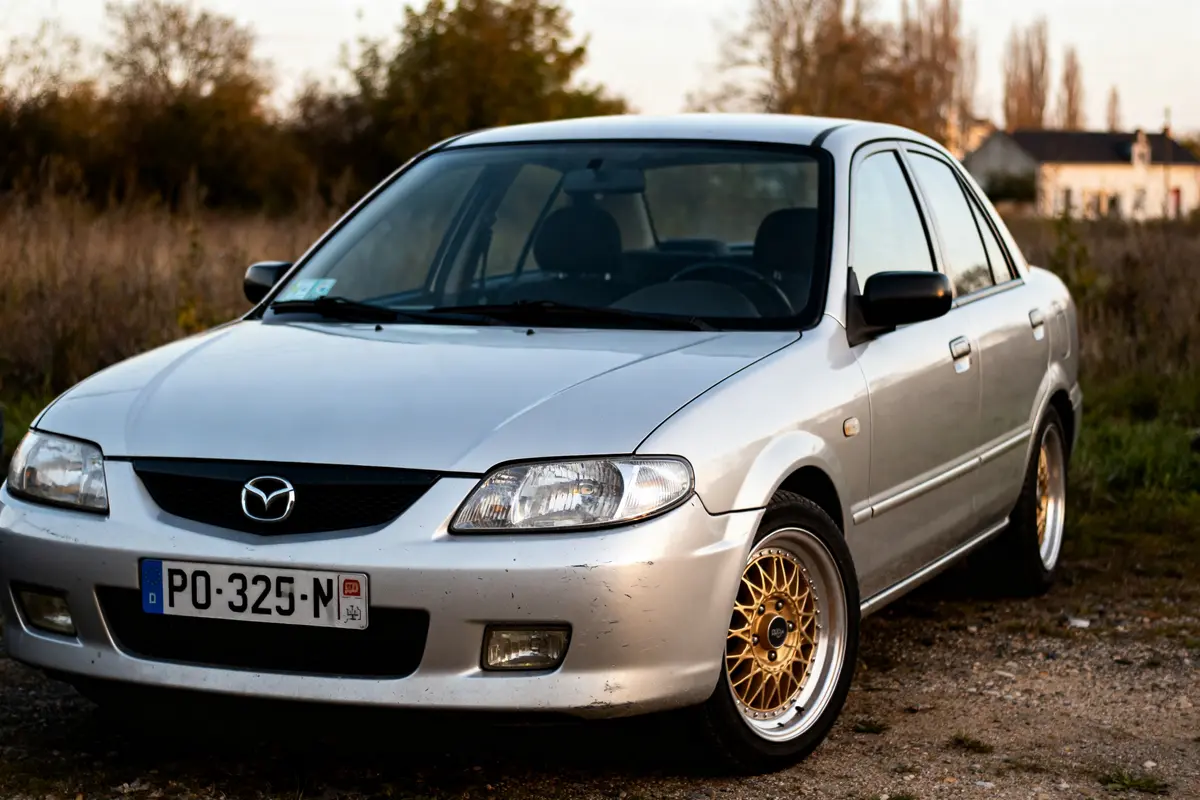 Voiture Mazda 323 Protegé argentée avec jantes dorées stationnée en extérieur rural