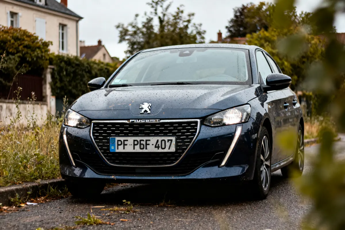 Voiture Peugeot 201 sombre garée sur route mouillée devant maisons et végétation