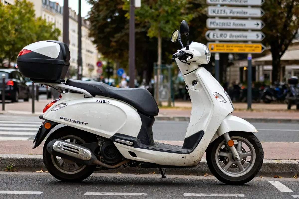 Scooter Peugeot 50CC blanc garé en ville devant un panneau de signalisation