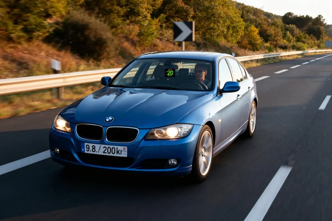 Voiture BMW bleue roulant sur autoroute bordée d'arbres, ciel dégagé, direction indiquée par panneau.
