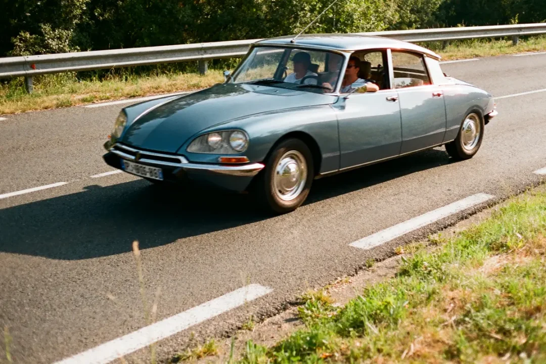 Voiture classique bleue roulant sur une route rurale ensoleillée, deux personnes à l'intérieur.