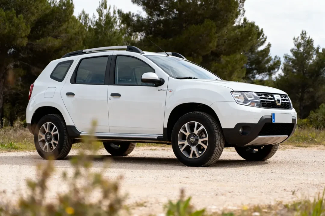 SUV blanc garé en extérieur, vue de profil, avec arrière-plan boisé et sol poussiéreux, ciel clair.