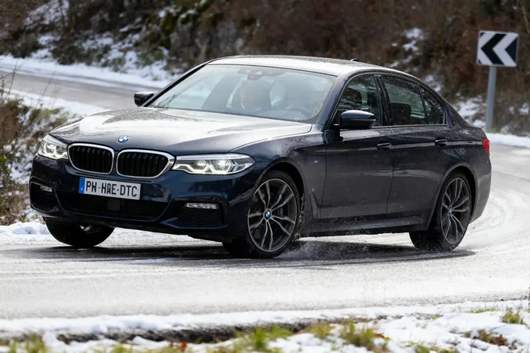 Voiture BMW noire roulant sur une route enneigée, virage serré à droite en hiver.