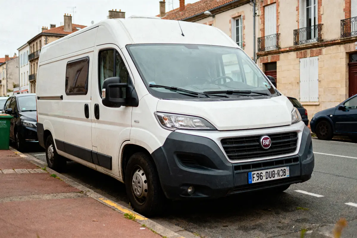Fourgon blanc Fiat garé en ville, bâtiments en brique en arrière-plan, voitures stationnées autour.