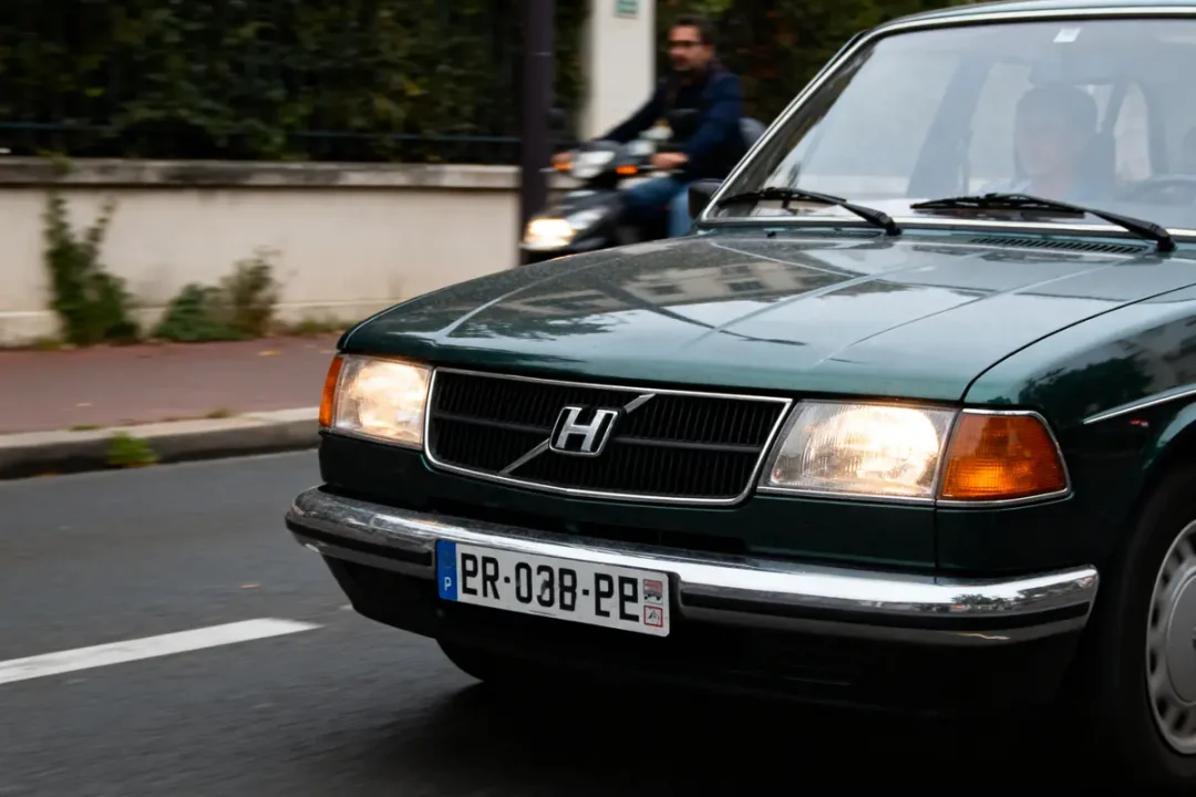 Voiture verte avec logo modifié, plaque d'immatriculation visible, moto floue en arrière-plan.