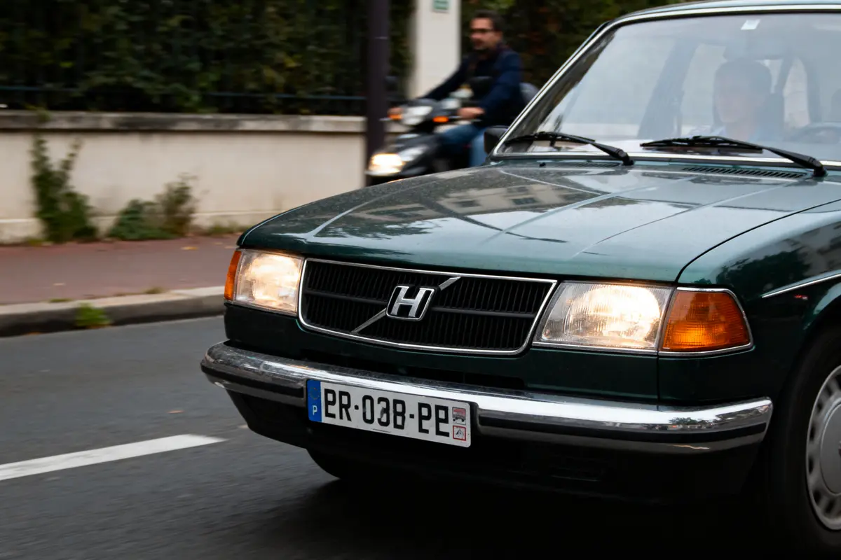 Voiture verte avec logo modifié, plaque d'immatriculation visible, moto floue en arrière-plan.