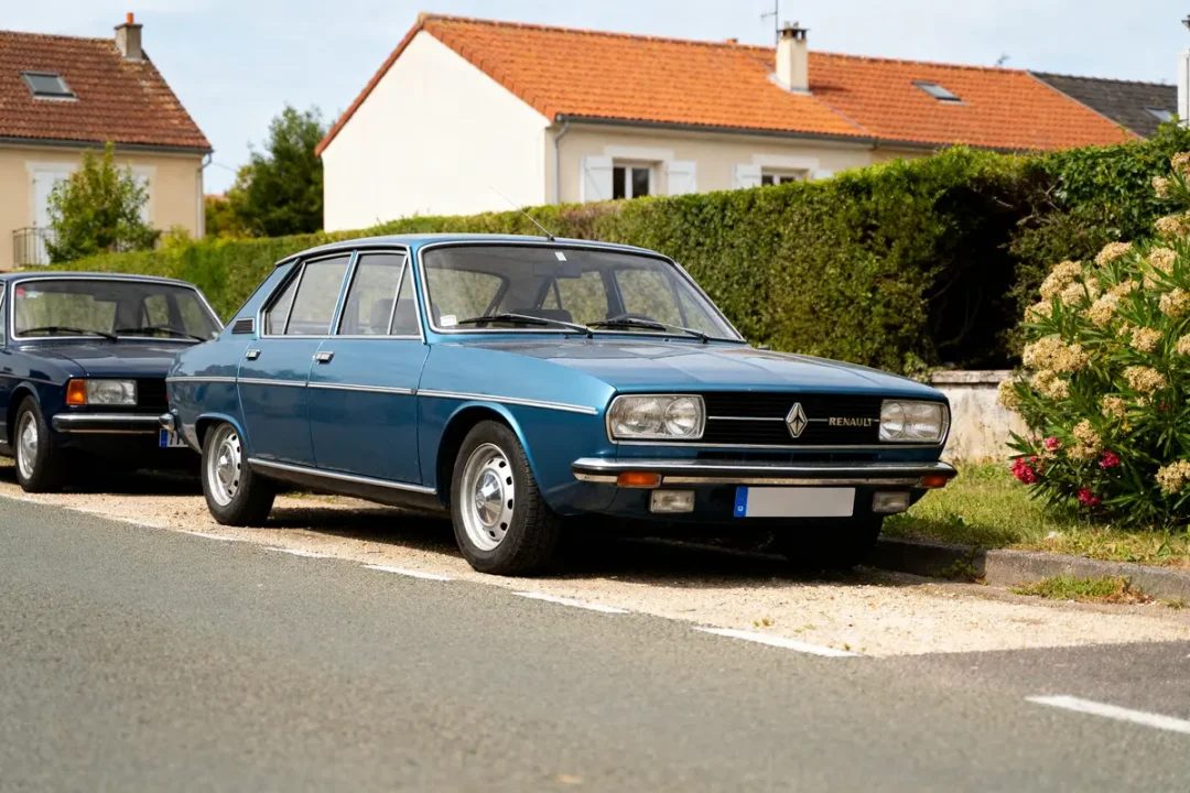 Berline classique Renault bleue garée devant une maison avec jardin, voiture stationnée derrière.