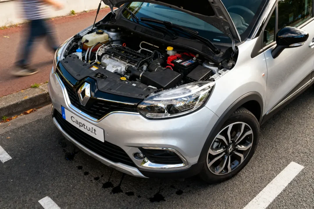 Renault Captur grise, capot ouvert montrant moteur, stationnée sur rue; piéton flou en mouvement.