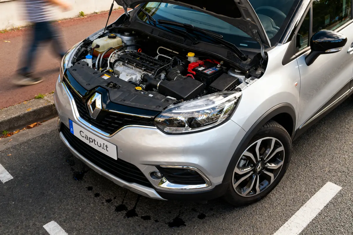 Renault Captur grise, capot ouvert montrant moteur, stationnée sur rue; piéton flou en mouvement.
