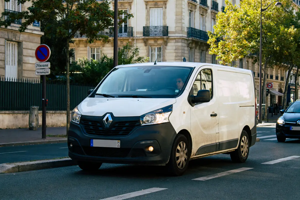 Fourgonnette Renault blanche en circulation urbaine, bâtiments haussmanniens et signalisation routière en arrière-plan.