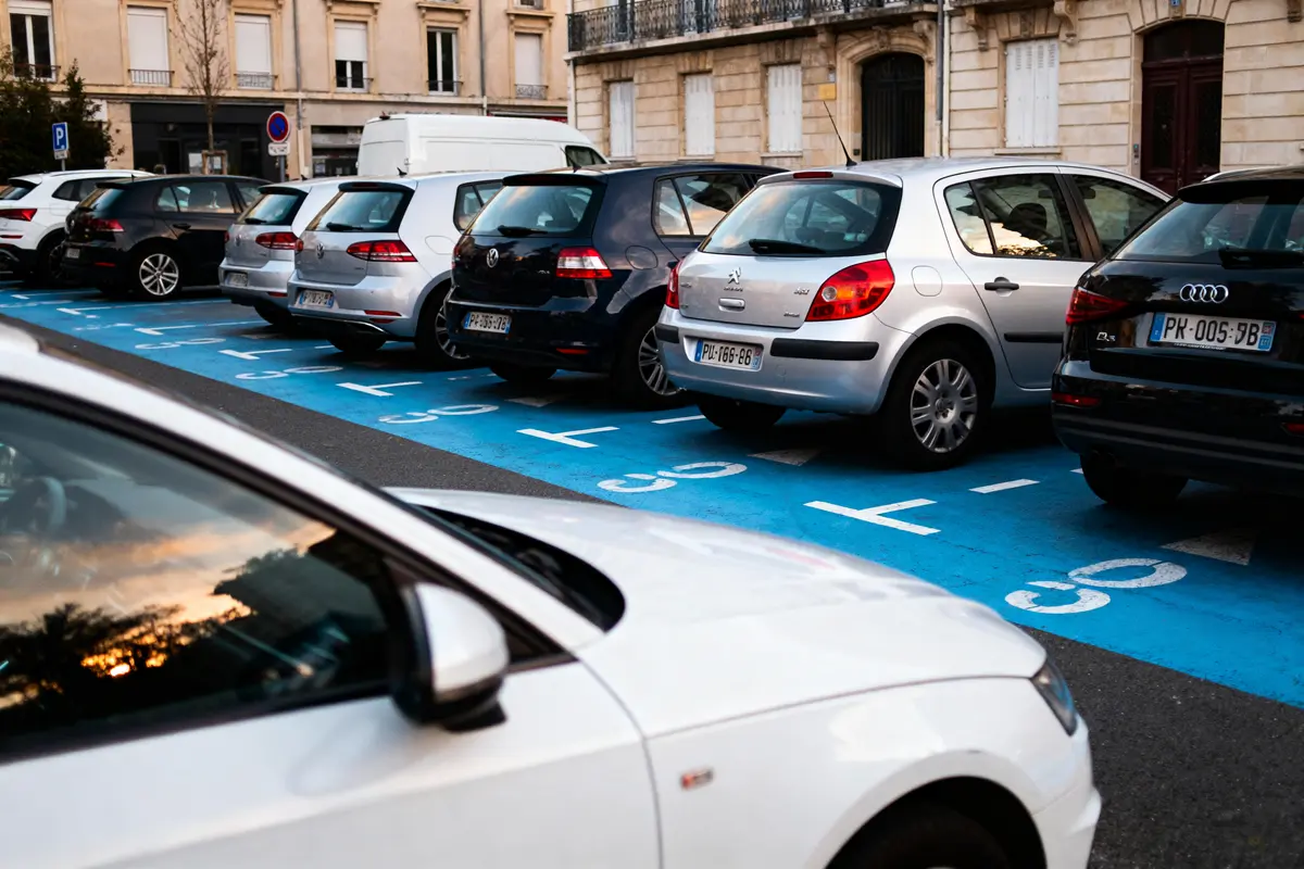Plusieurs voitures garées sur une aire de stationnement bleue en ville, bâtiments en arrière-plan.